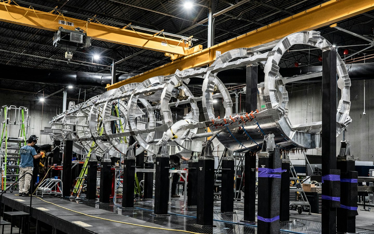 Two workers assemble a large metal aircraft frame in a factory.